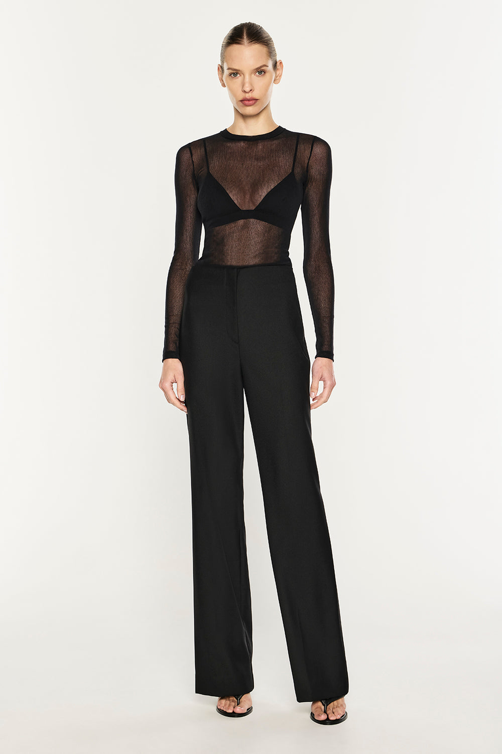 A model wearing a black sheer long-sleeve top with a black underwire bra insert and high-waisted black wide-leg trousers, photographed against a plain white studio background.