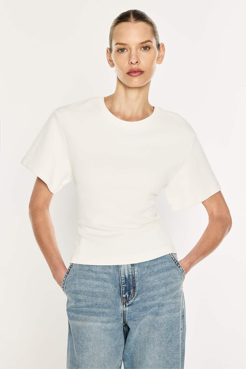 A model wearing a white short-sleeve crewneck top and light blue jeans, standing against a plain light gray studio background.