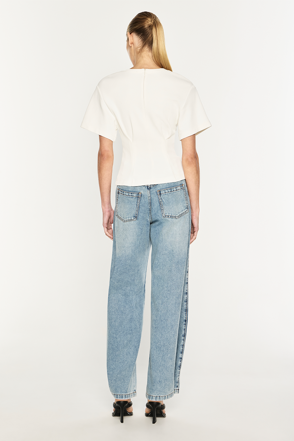 A woman shown from the back wearing a white short-sleeve top and light blue wide-leg jeans with side slits, paired with black heels, against a plain white studio background.