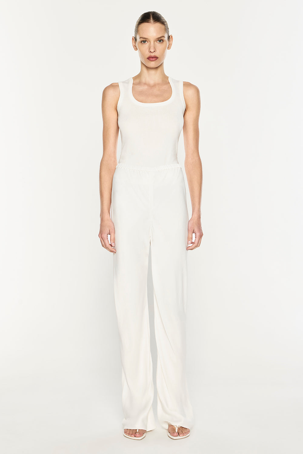A model stands against a plain white studio background wearing a white sleeveless jumpsuit with a wide scoop neckline and very wide-leg pants.
