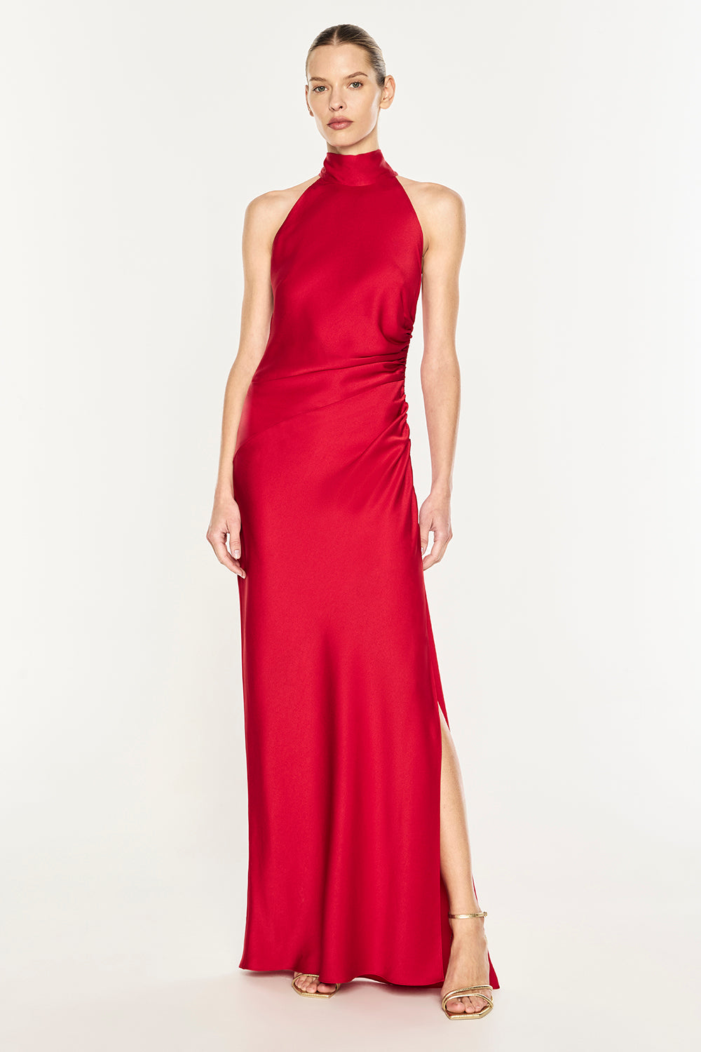 A woman modeling a sleeveless, high-neck, floor-length red gown with a side slit, photographed against a plain light studio background.