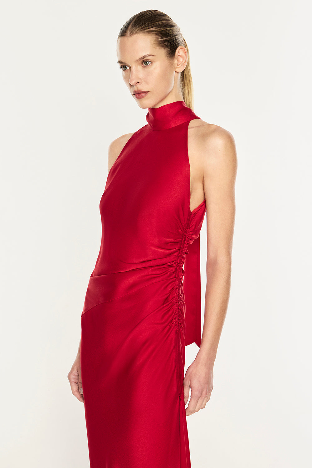 A model wearing a sleeveless, high-neck red dress with ruched detailing on the side, photographed against a clean white studio background.