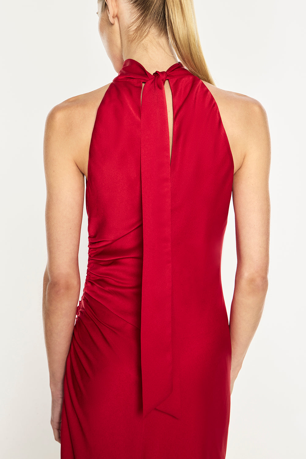 A woman shown from the back wearing a sleeveless red dress with a halter-style neckline, long tie detail at the back, and gathered draping fabric, photographed against a plain light background.