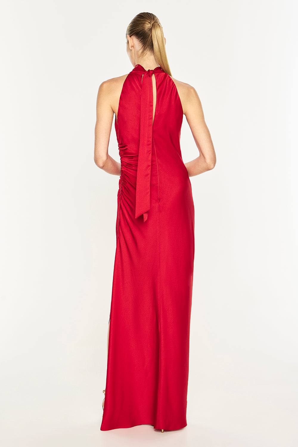 A woman viewed from the back wearing a bright red sleeveless halter-neck maxi dress with a gathered waist and long tie detail, photographed against a plain light background.