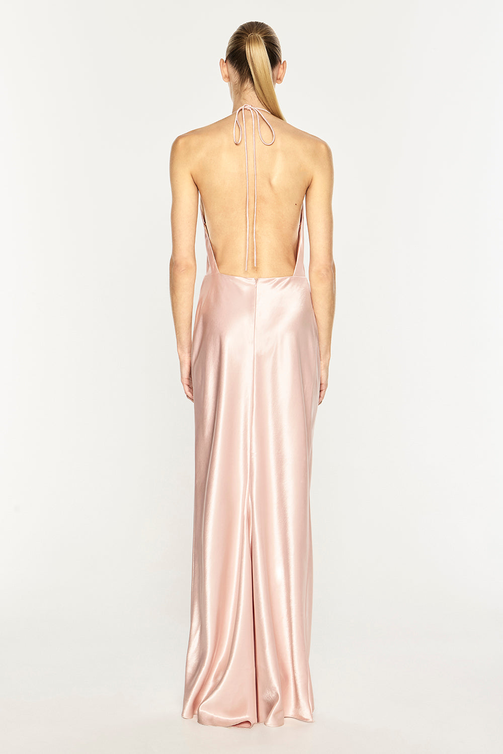 A woman viewed from the back wearing a floor-length satin gown in a pale blush pink color, with a tie-back neckline and an open back, against a plain light gray studio background.