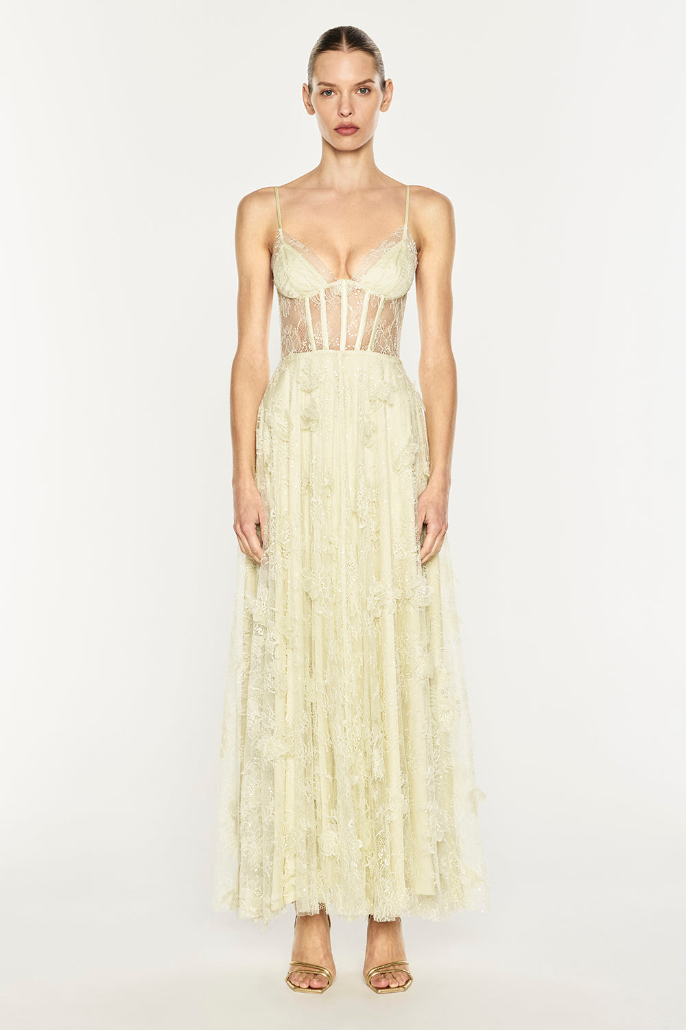 A model wearing a pale cream or ivory lace and tulle maxi dress with a sheer corset-style bodice and thin straps, standing against a plain light gray studio background.
