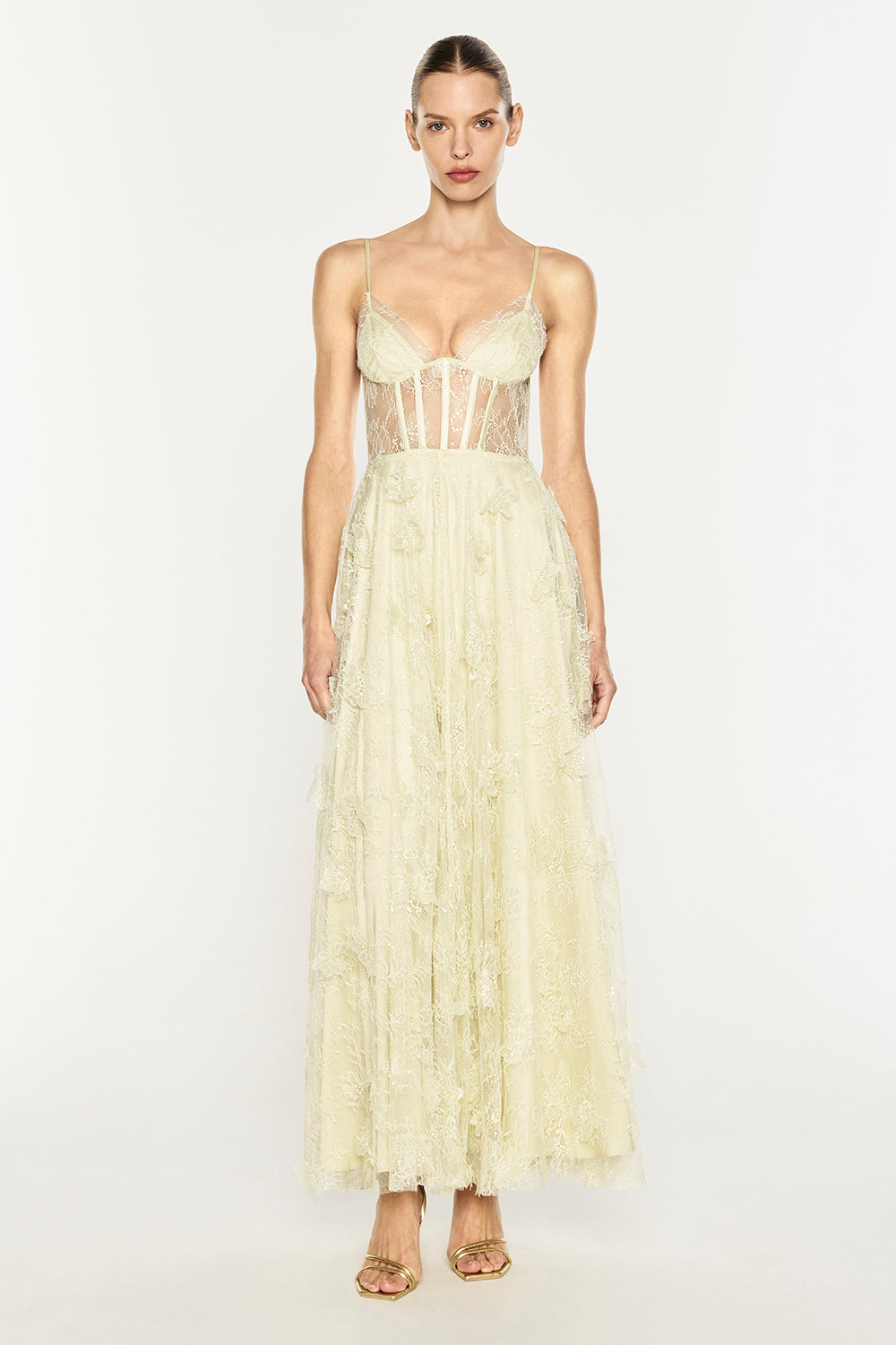 A model wearing a pale yellowish-cream lace maxi dress with a sheer corset-style bodice and a long, layered tulle skirt, standing against a plain white studio background.