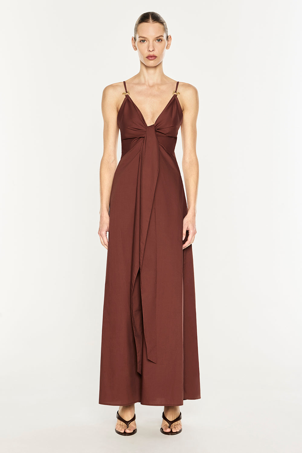 A woman modeling a floor-length rust-brown maxi dress with thin spaghetti straps, a deep V-neckline, and a gathered waist detail, set against a plain light gray studio background.