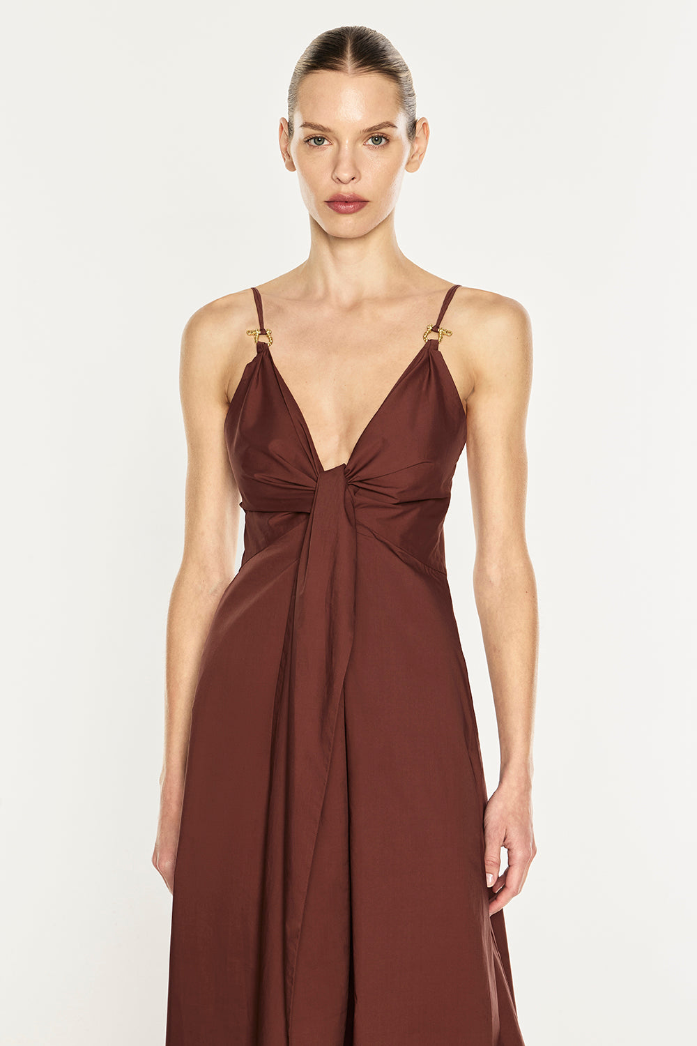 A woman modeling a sleeveless, deep V-neck maxi dress in a rich rust brown color, featuring thin straps with gold-toned hardware and a gathered front detail, set against a plain light gray studio background.