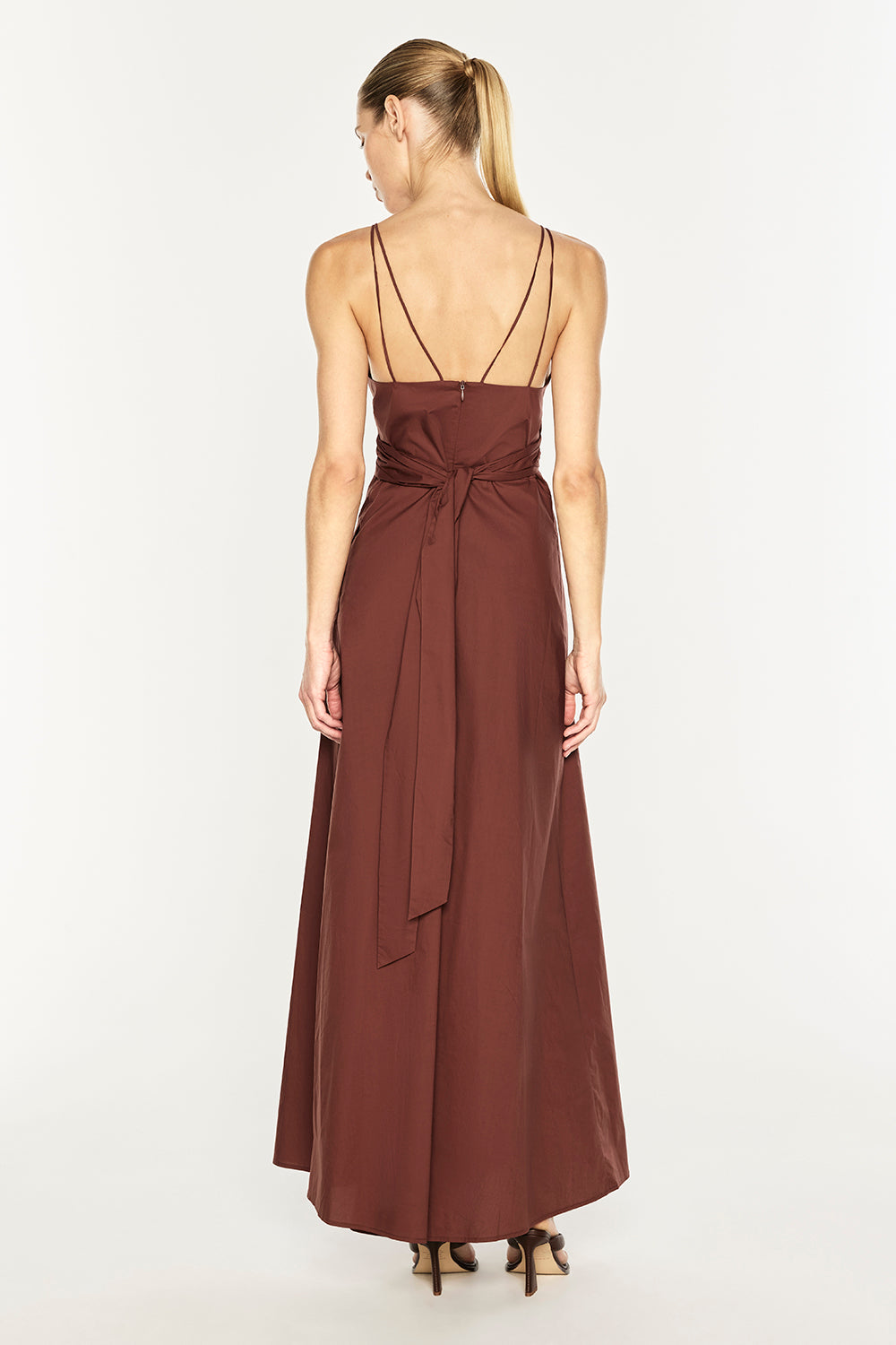 A woman wearing a long, sleeveless rust-brown maxi dress with thin double straps and a tied waist, shown from the back against a plain white studio background.