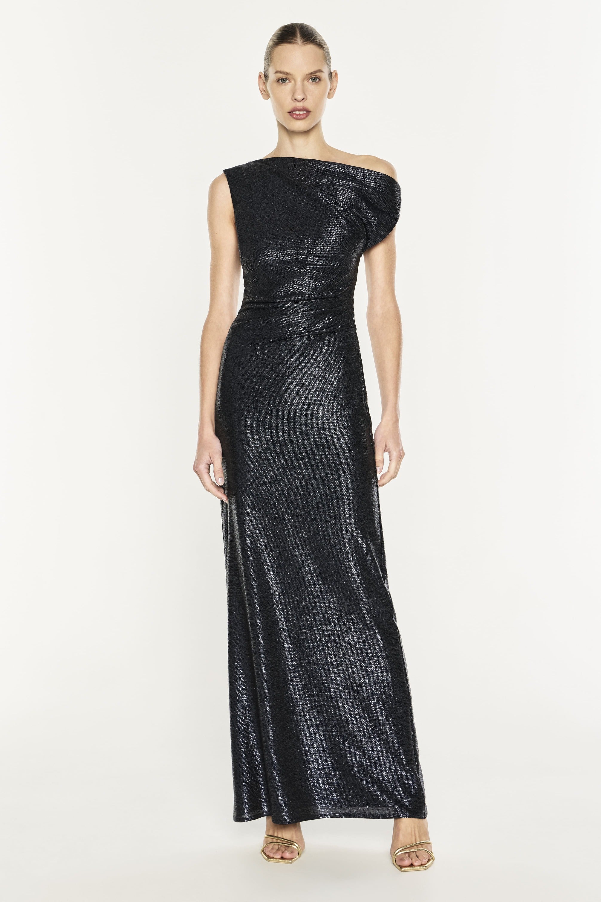 A model wearing a floor-length, off-the-shoulder black dress with a textured, shimmering finish, paired with metallic gold high-heeled sandals, set against a plain white studio background.