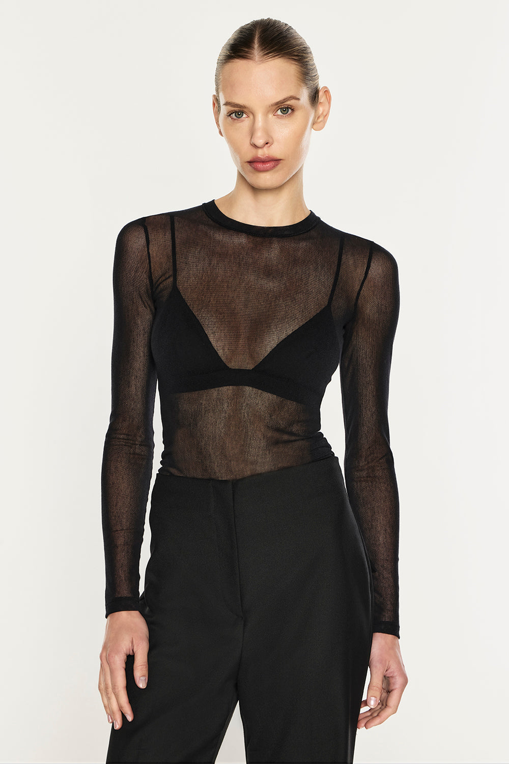 A model wearing a sheer black long-sleeve mesh top with a black underwire bra and black trousers, photographed against a light gray studio background.