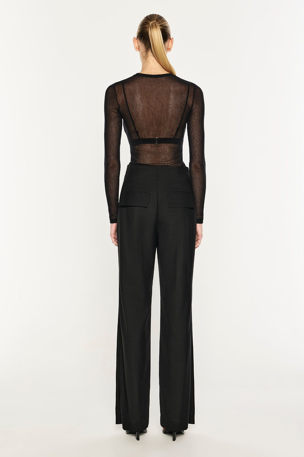 A model viewed from the back wearing a black sheer long-sleeve top with a wide waistband and black wide-leg trousers, against a plain white studio background.