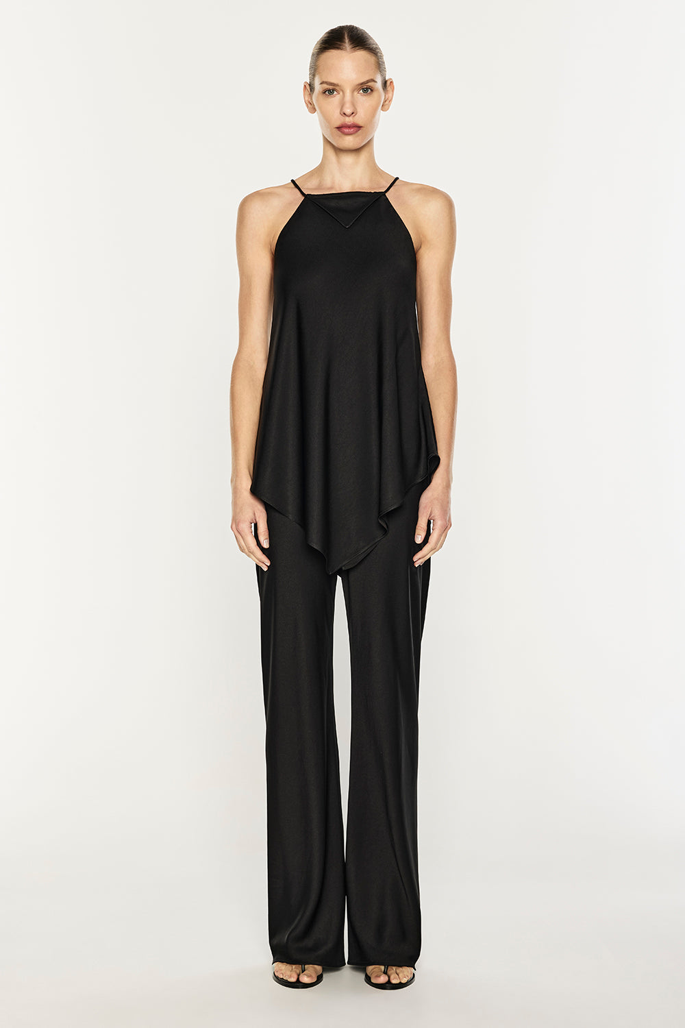 A model stands against a plain light gray studio background wearing a black sleeveless draped top with matching wide-leg pants and black sandals.