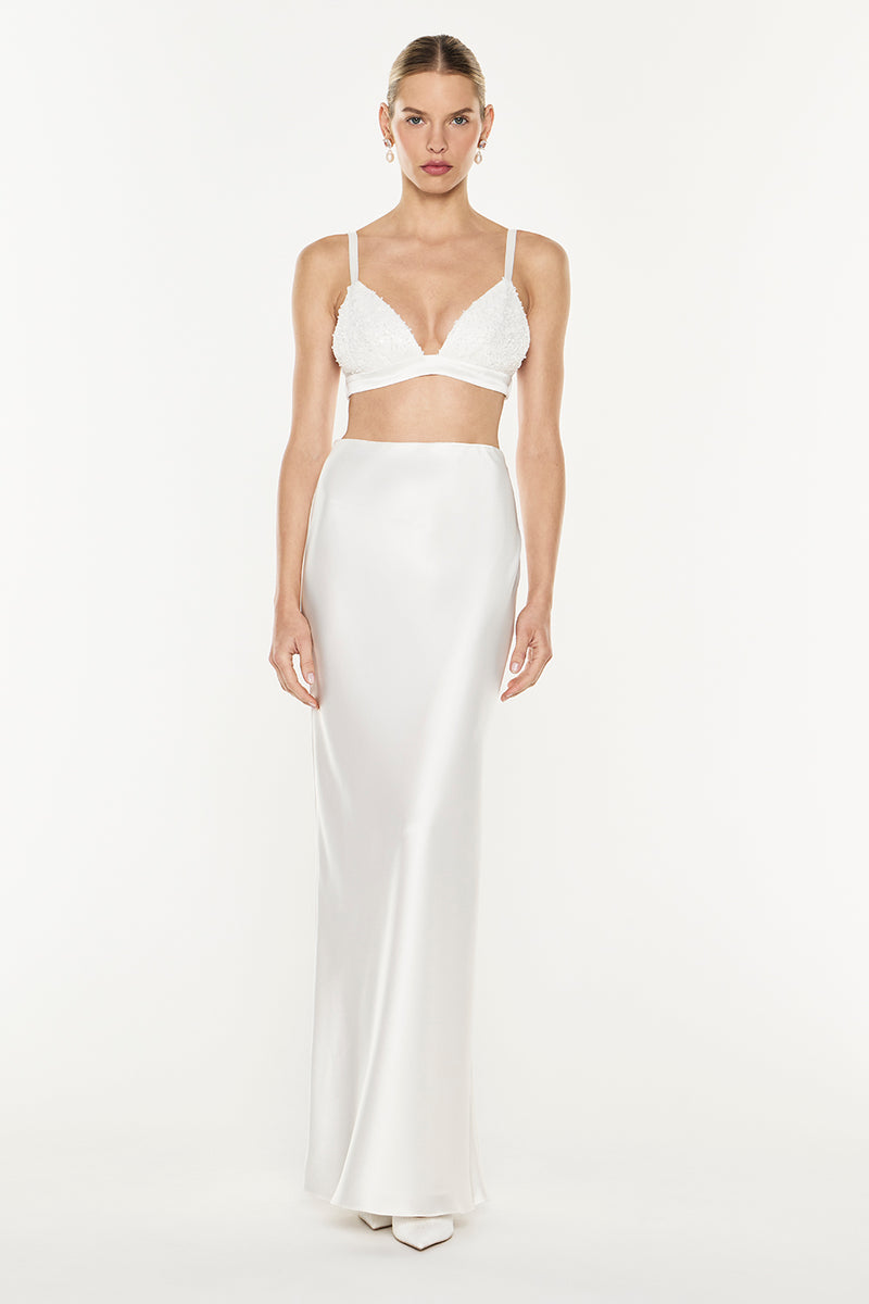 A model wearing a white two-piece formal outfit consisting of a lace bustier crop top with thin straps and a matching satin maxi skirt, shot against a seamless white studio background.