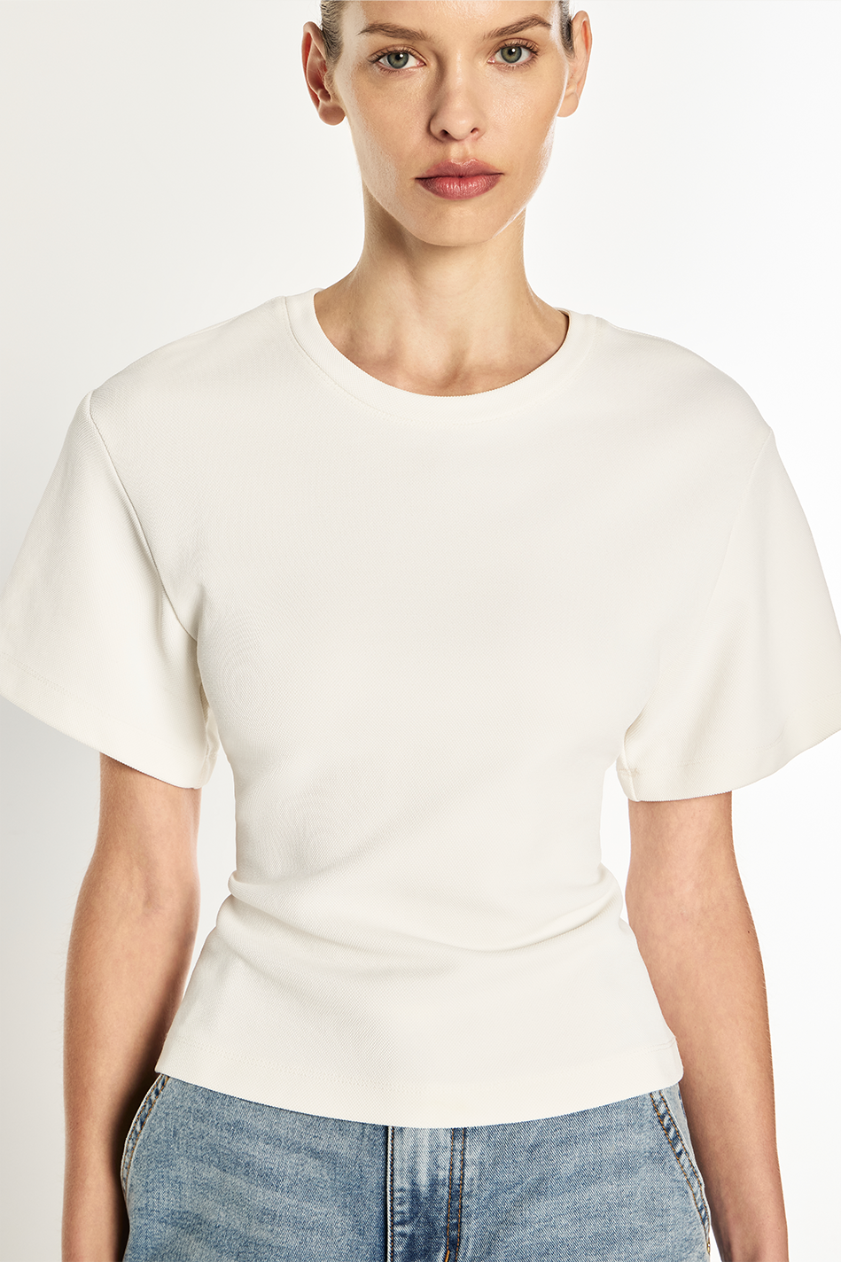 A model wearing a cream-colored short-sleeve crewneck top and light blue jeans, photographed against a clean white studio background.