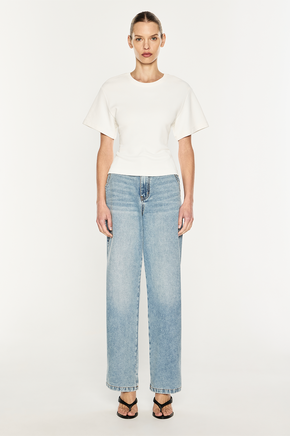 A model stands against a plain light background wearing a white short-sleeve top, light-wash wide-leg blue jeans, and black sandals.