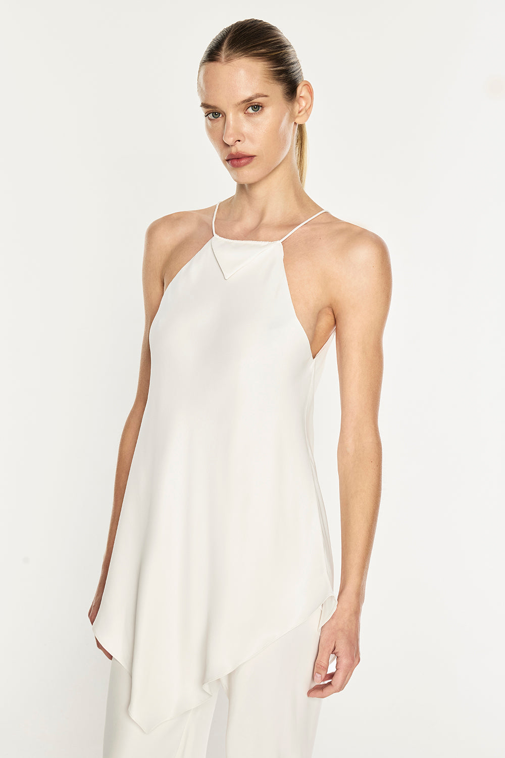 A woman modeling a white sleeveless halter top with a draped, asymmetrical hem against a plain white studio background.