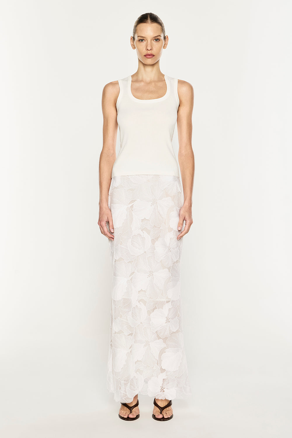 A model stands against a plain white studio background wearing a Manning Cartell sleeveless white ribbed tank top and a long white sheer floral lace skirt, paired with black strappy sandals.