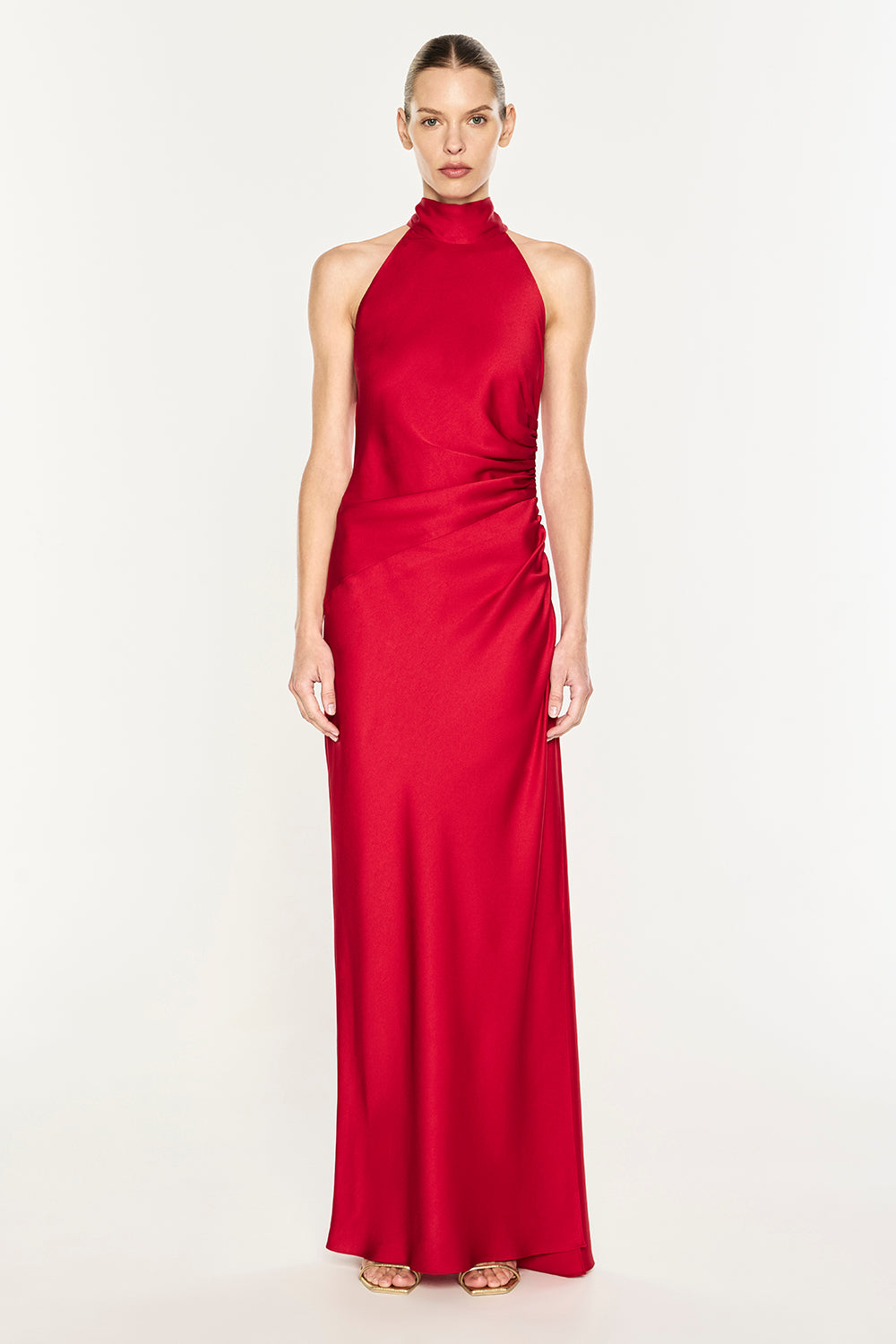 A model wearing a vibrant red halter-neck maxi dress with a draped, gathered waist, photographed against a seamless white studio background.