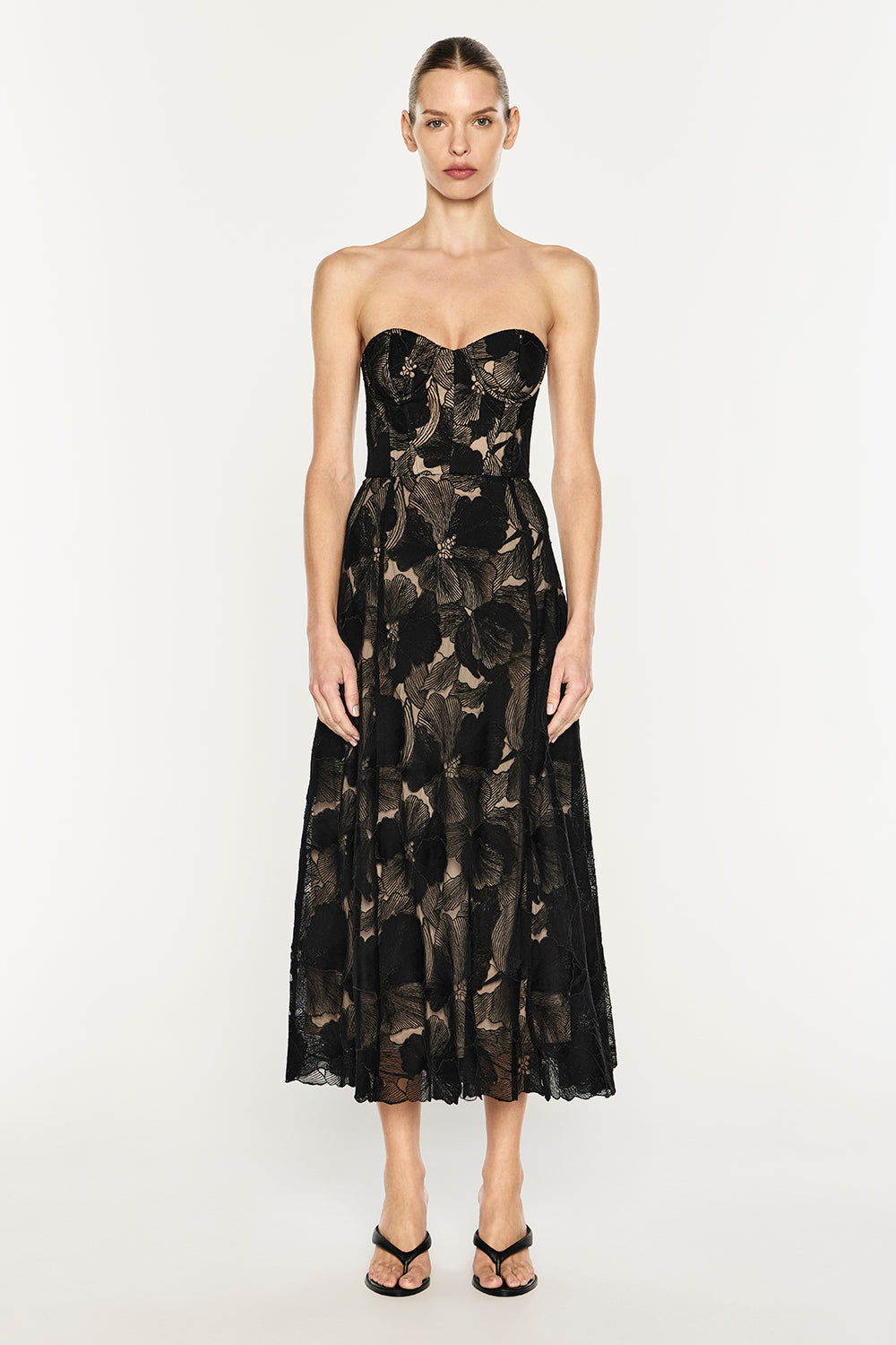 A woman modeling a strapless black midi dress with a sheer floral lace overlay and a flared skirt, paired with black strappy heels, against a plain light gray studio background.