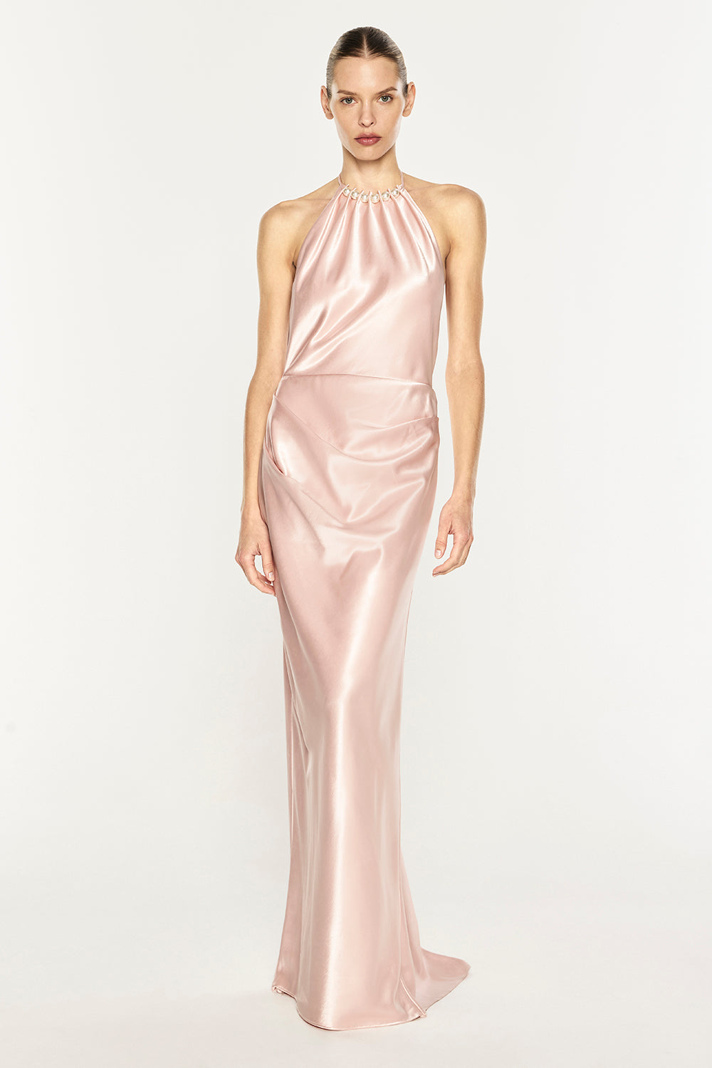 A model wearing a floor-length, satin-like halter gown in a soft blush pink color, standing against a plain white studio background.