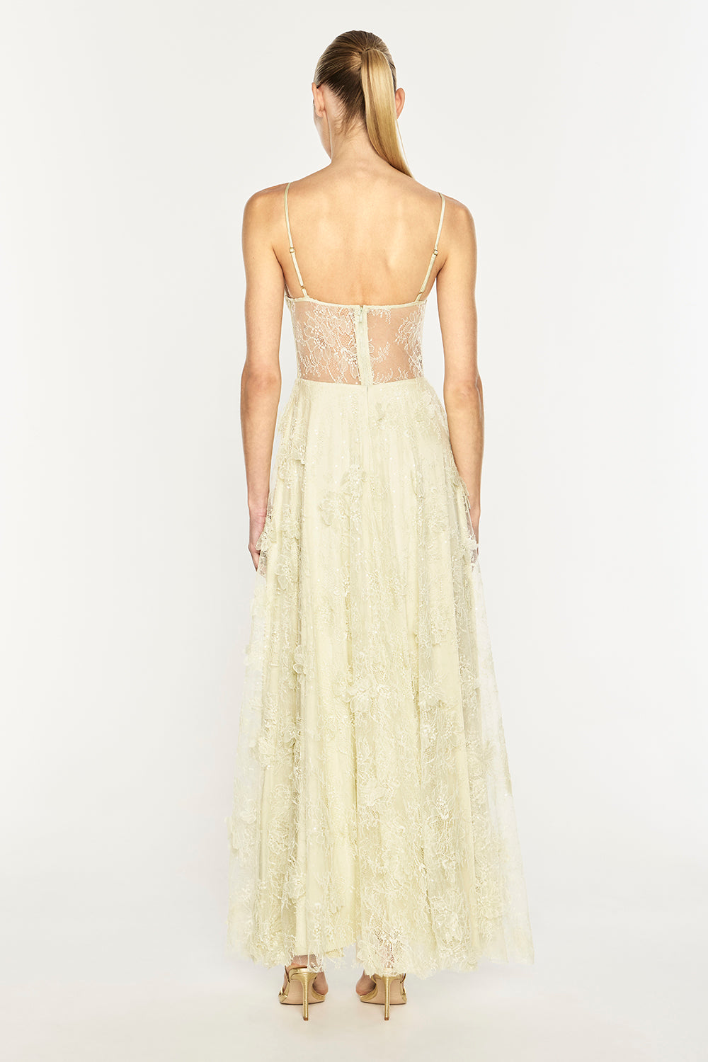 A woman viewed from the back wearing a long cream lace dress with thin straps, a sheer lace bodice, and a full layered tulle skirt, standing against a plain light background.