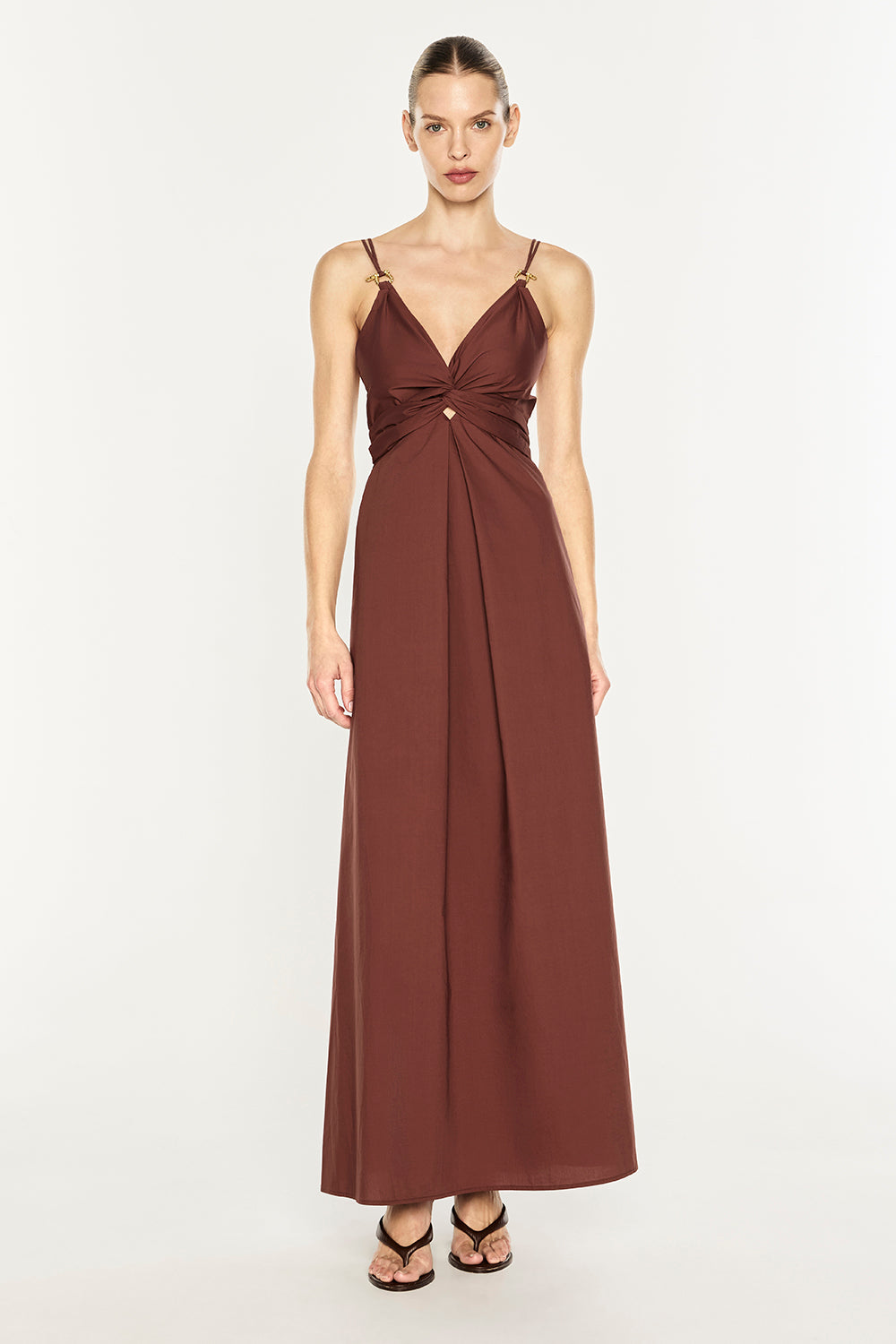 A woman modeling a floor-length brown maxi dress with thin straps, a V-neckline, and a gathered twist detail at the waist, photographed against a plain light background.