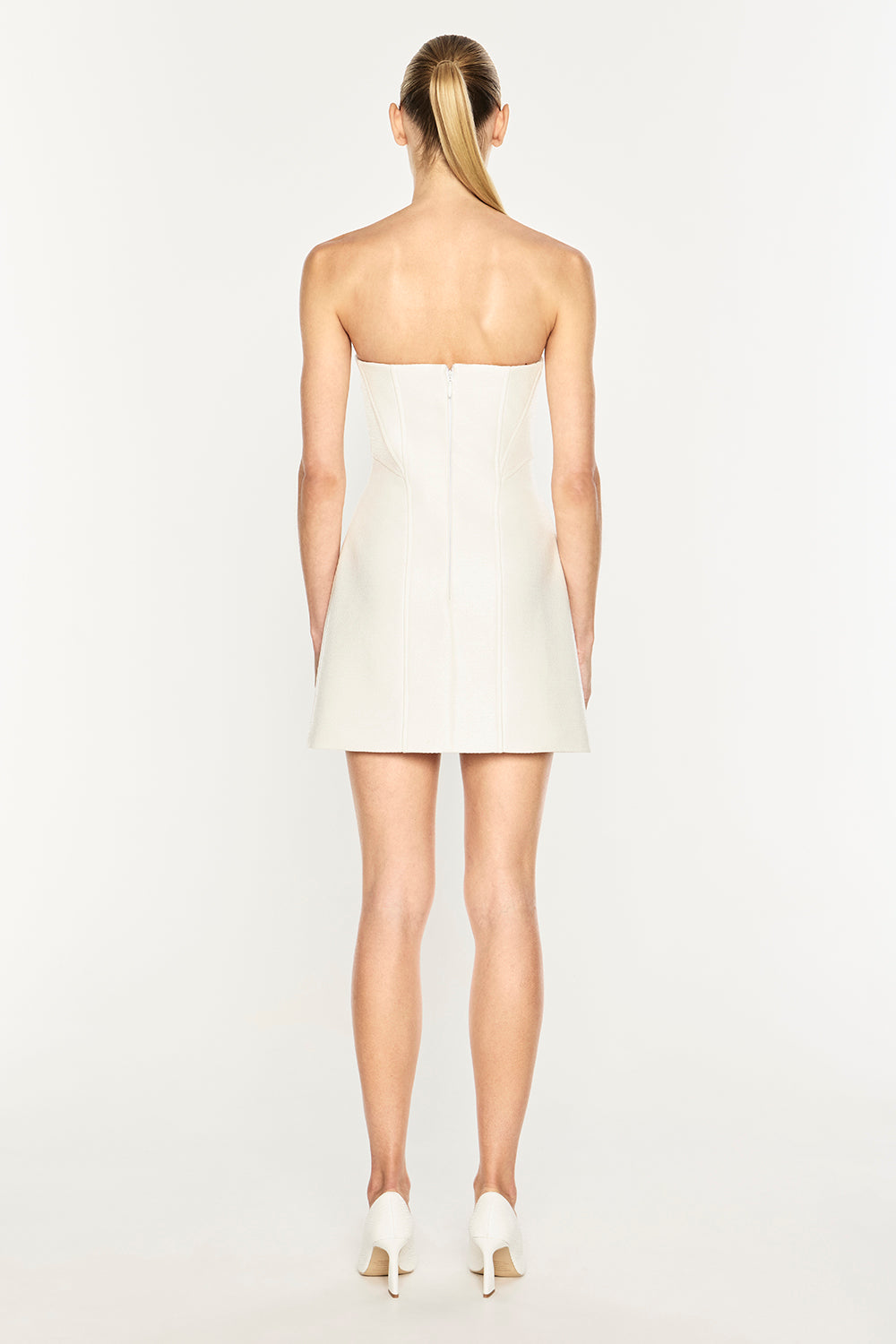 A woman shown from the back wearing a strapless off-white mini dress and white heels against a plain light gray studio background.